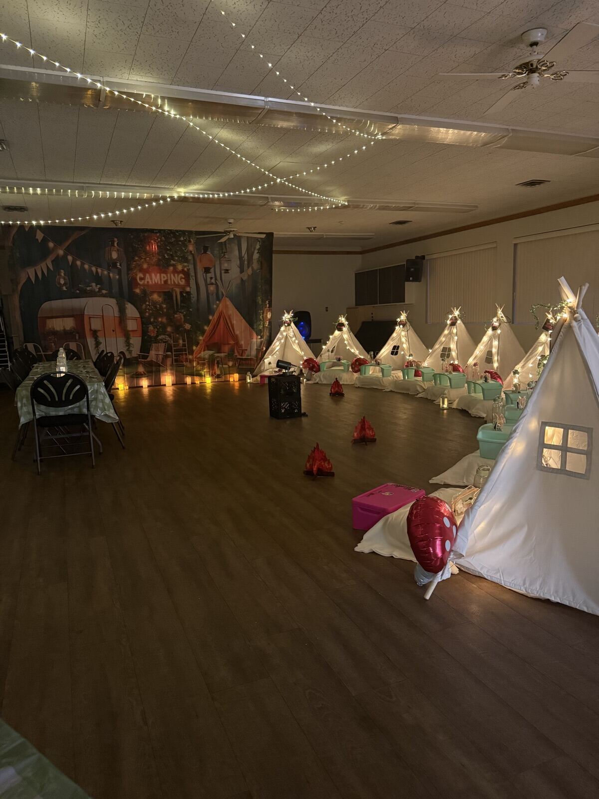 A row of white teepees under string lights in the dimmed main hall for a birthday sleepover